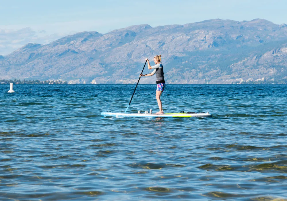 A woman enjoys paddle boarding on serene waters, with stunning mountains rising in the background.
