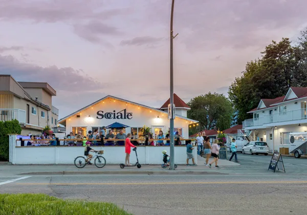 Children are riding bikes and a scooter in front of the Socialē eatery's busy patio on a beautiful evening.