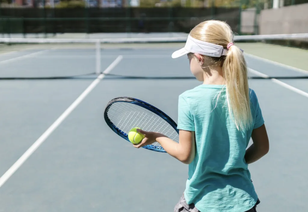 A young girl stands confidently with a tennis racket in one hand and a ball in the other, eager to hit the court.